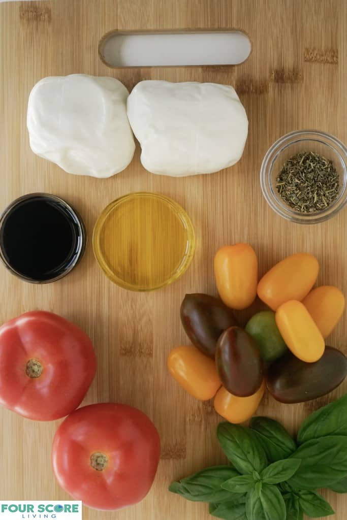 Close up aerial view of the ingredients to make burrata caprese, including small clear glass bowls with dried herbs, oil ,and balsamic vinegar, two sprigs of fresh basil, two balls of burrata cheese, two red tomatoes and yellow, purple and green plum tomatoes on a cutting board with a sharp knife.