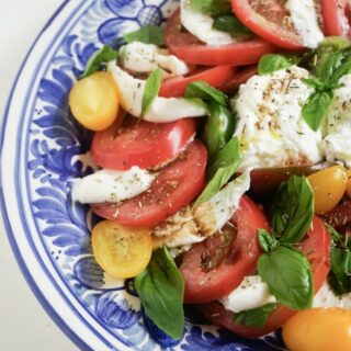 Up close, aerial, partial view of burrata caprese with burrata cheese, sliced red tomatoes, sliced yellow Roma tomatoes, fresh basil, drizzled with oil and balsamic vinegar and sprinkled with fresh herbs on a blue plate.