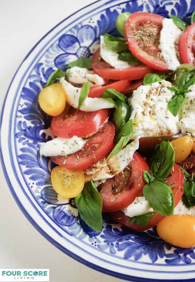 Up close, aerial, partial view of burrata caprese with burrata cheese, sliced red tomatoes, sliced yellow Roma tomatoes, fresh basil, drizzled with oil and balsamic vinegar and sprinkled with fresh herbs on a blue plate.