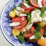 A burrata caprese with burrata cheese, sliced red tomatoes, sliced yellow Roma tomatoes, fresh basil, drizzled with oil and balsamic vinegar and sprinkled with fresh herbs on a blue plate.