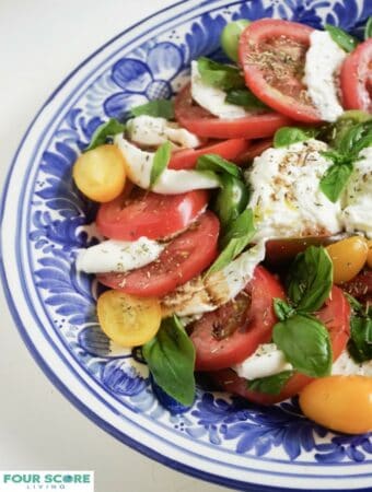 Up close, aerial, partial view of burrata caprese with burrata cheese, sliced red tomatoes, sliced yellow Roma tomatoes, fresh basil, drizzled with oil and balsamic vinegar and sprinkled with fresh herbs on a blue plate.