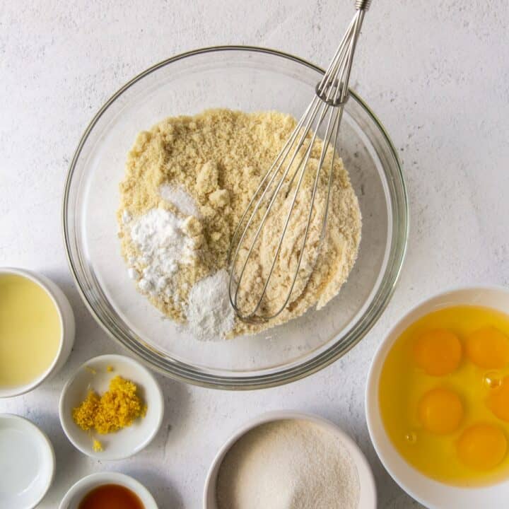 Glass bowl with dry keto lemon cake ingredients being whisked together. Individual bowls with the wet ingredients are also on the countertop.