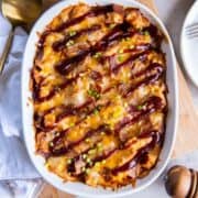 Oval casserole dish with a baked BBQ chicken casserole drizzled with BBQ sauce and set on a wooden cutting board next to a kitchen towel and a large round serving spoon.