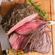 An herb crusted, juicy and tender beef bottom round roast partially sliced on a wood cutting board with a spring of fresh rosemary and a two pronged serving tool and a sharp knife.